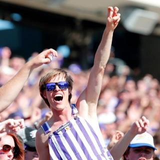 一个快乐的, 在一场拥挤的足球比赛中，一名身穿条纹工作服、戴着紫色太阳镜的欢呼学生做出了两个手指的“青蛙加油”手势.