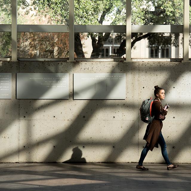 一名学生背着双肩包穿过Moudy大楼，建筑特色在混凝土墙上投下了戏剧性的阴影